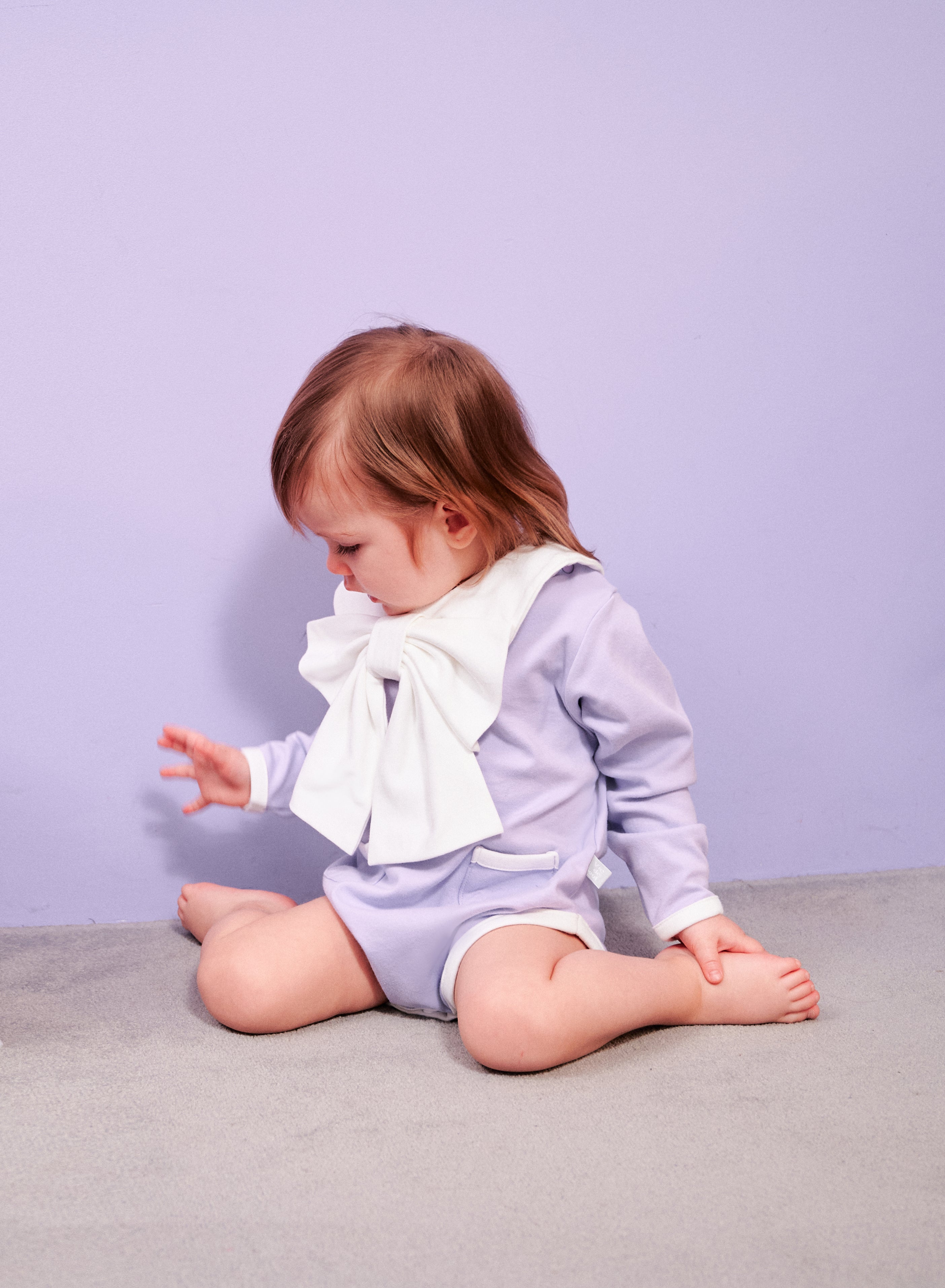 Dress Up Bodysuit (Lavender): Minimalistic design with contrasting lines and charming pocket details. Paired with a ribbon scarf bib for a formal look.